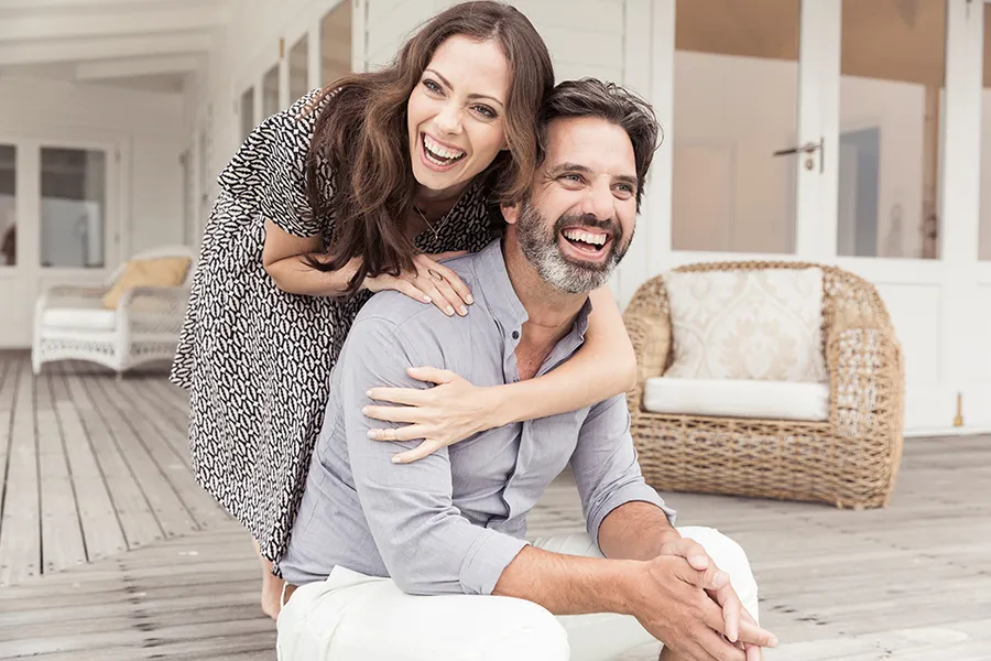 Health-and-Wellness-Services-Doctor A middle-aged couple on a wooden porch with wicker furniture; the woman is playfully hugging the man from behind. They are benefiting from the health and wellness services from Genifer Chavez, MD in Tucson.